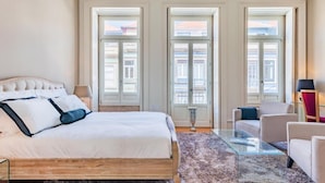 Habitación de hotel con una cama grande, una mesita de centro de vidrio, un sofá y vistas a edificios a través de las ventanas.