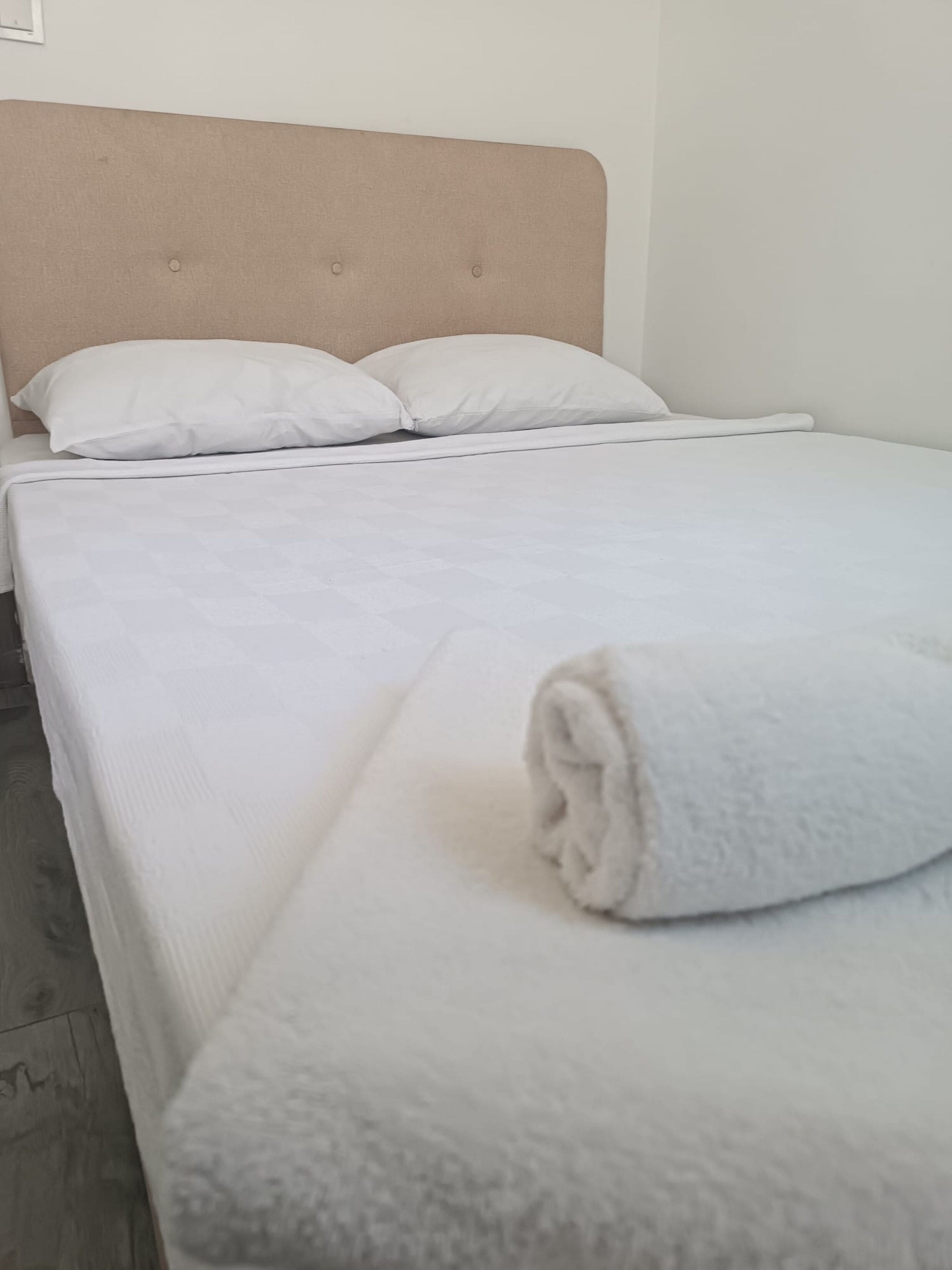 A bed with a beige headboard, white bedding, and a rolled towel on the footboard.