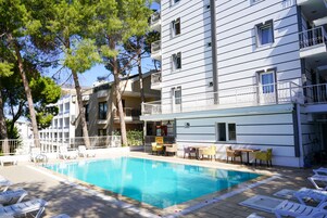 Una piscina con sillones de descanso y zona de bar, rodeada por un edificio de varios pisos con balcones.