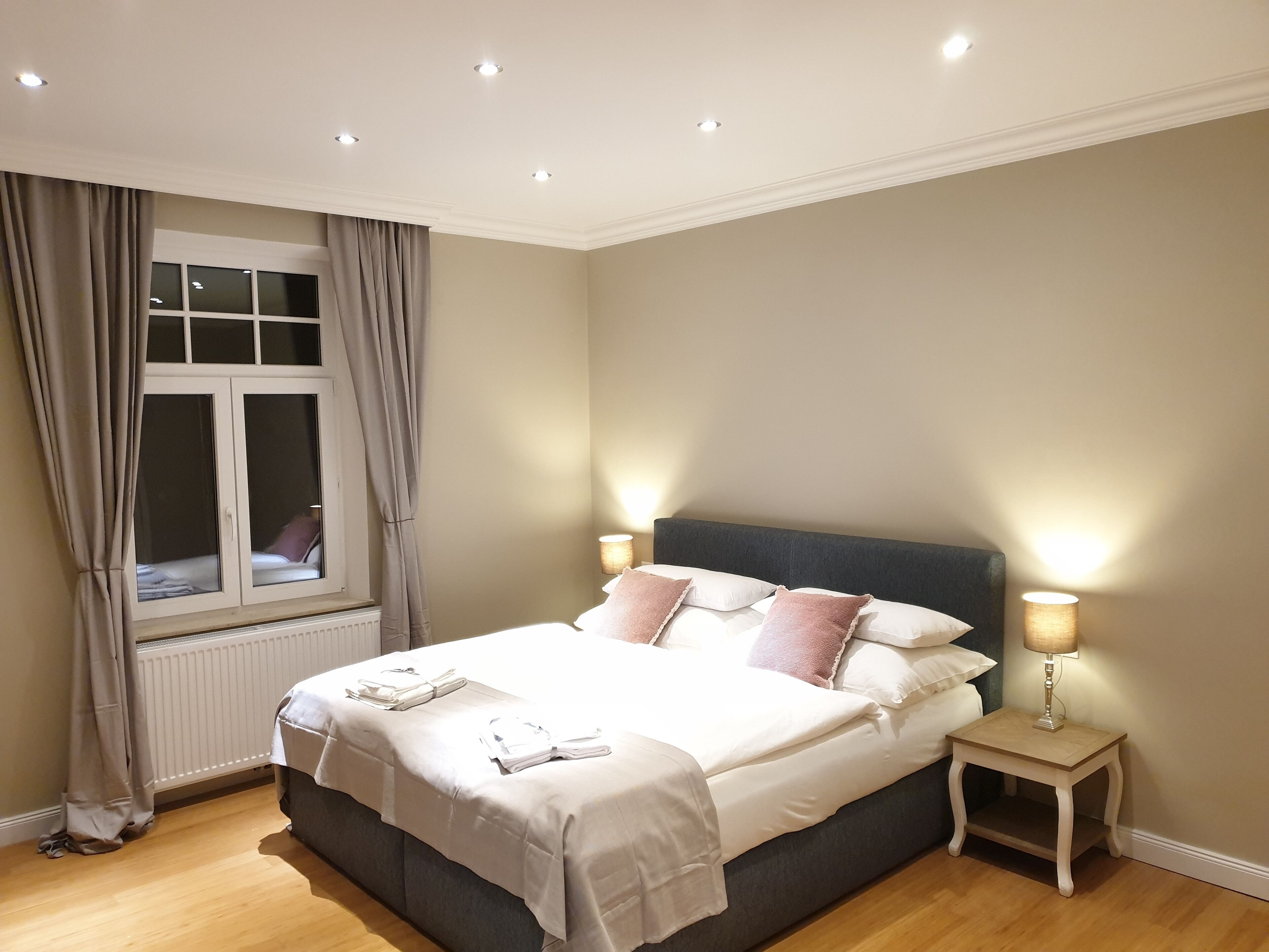 Una cama bien tendida con almohadas y una colcha, una mesita de noche con lámpara, una ventana con cortinas e iluminación empotrada en el techo.