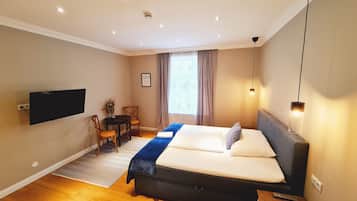 Ein Schlafzimmer mit Bett, einem an der Wand befestigten Fernseher, einem Schreibtisch mit Stuhl und einem Fenster mit Vorhängen.