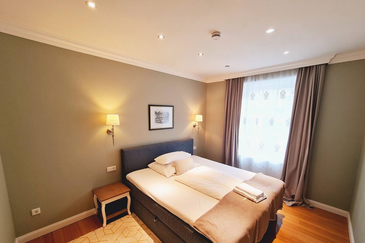 A neatly arranged bedroom with a large bed, two bedside lamps, a small bedside table, and a window with curtains.