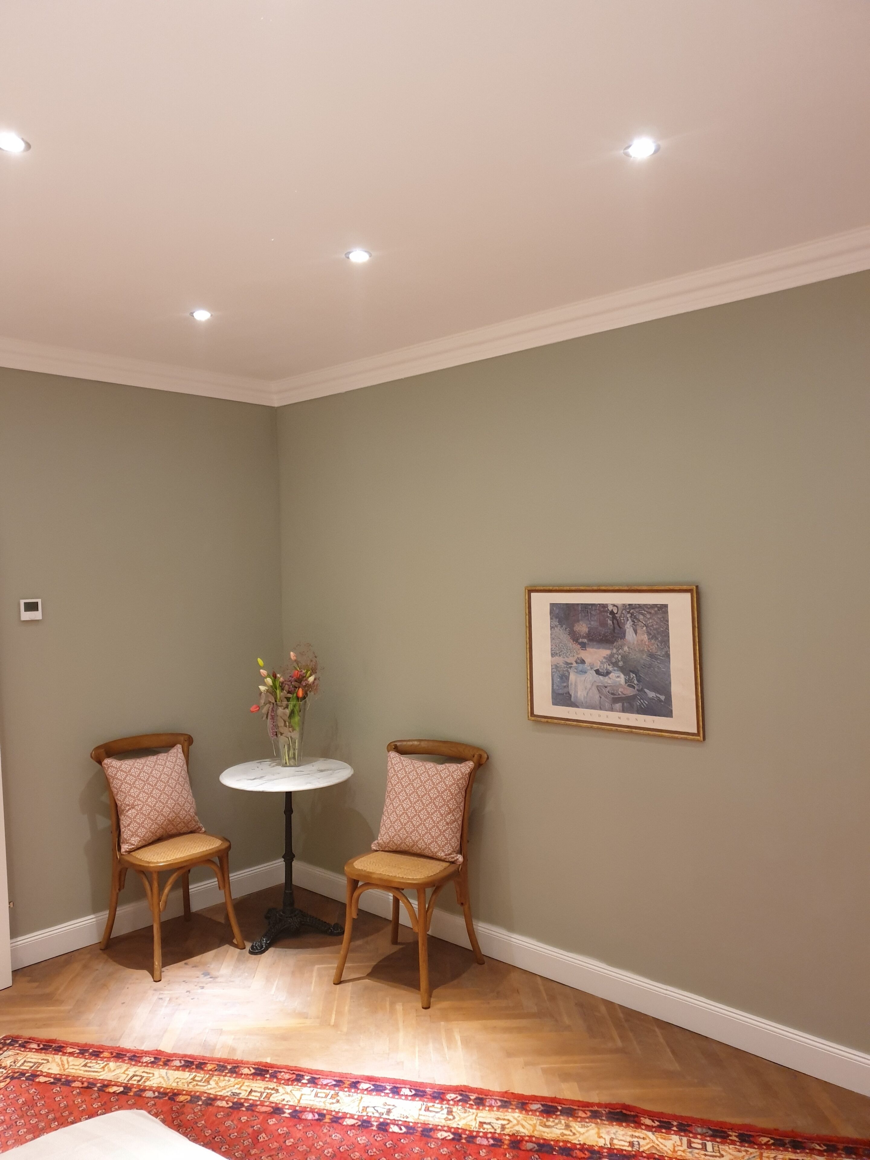 Una habitación con dos sillas de madera, una mesita redonda con un florero y un cuadro en la pared, además de un tapete estampado en el suelo.