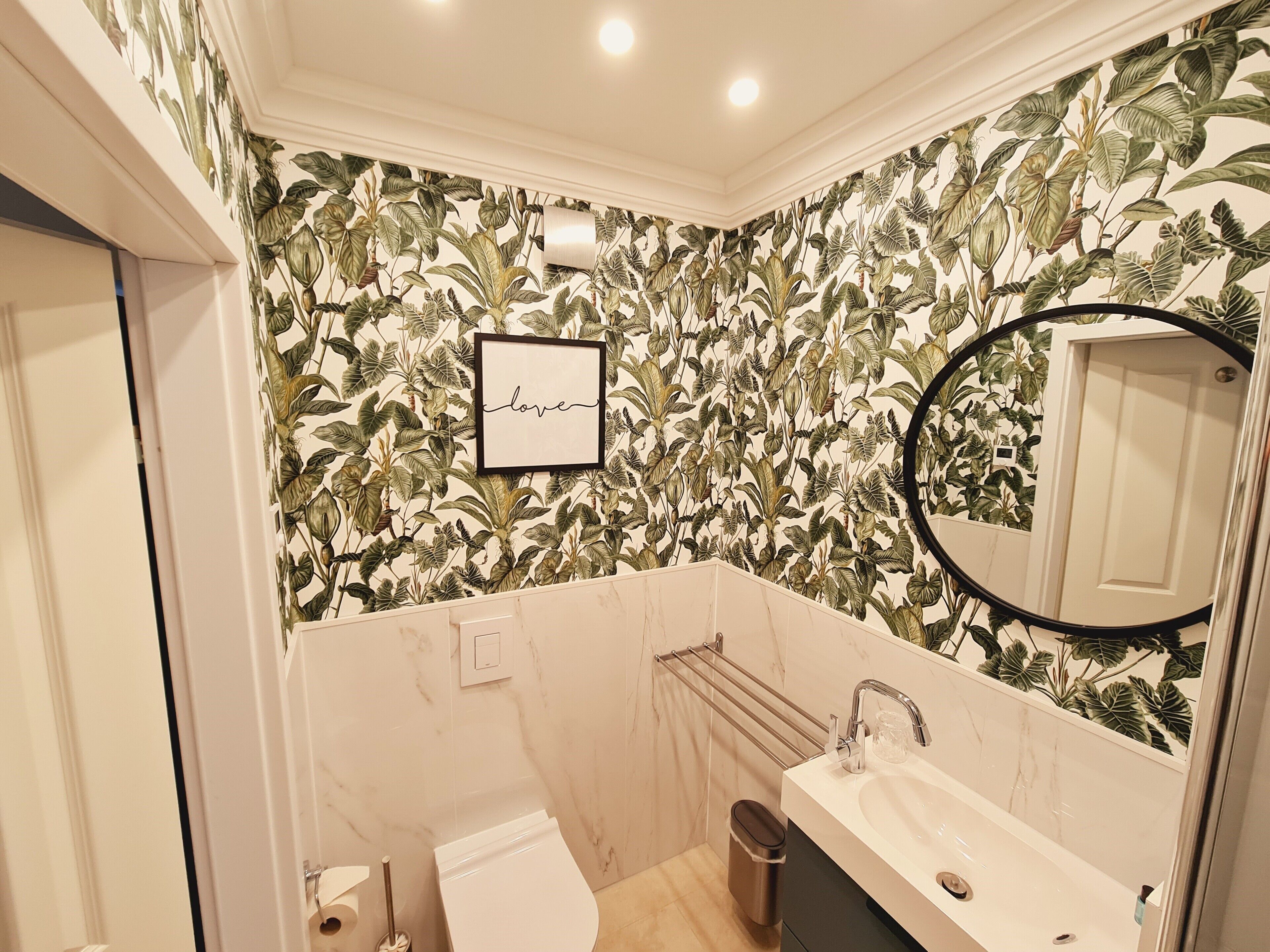 Un baño con papel tapiz floral, un cartel enmarcado que dice 'love', un espejo y un lavamanos moderno.