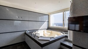 A modern bathroom with a large bathtub, a flat-screen TV, and a view of the city through a large window. - Radisson Blu Erfurt (Erfurt)
