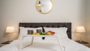 Cama bem arrumada com lençóis brancos, um tabuleiro com frutas e uma xícara, e duas luminárias de cabeceira de cada lado.