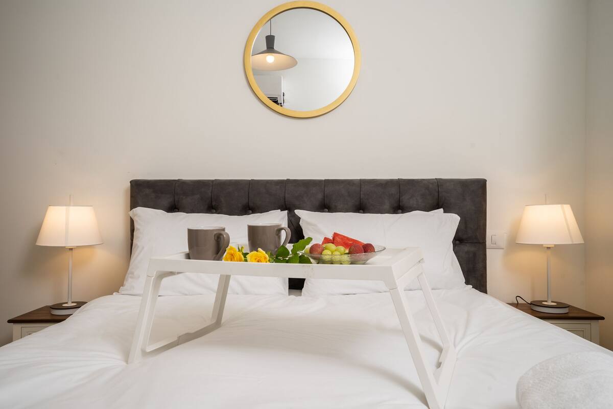 Un lit bien fait, avec du linge de lit blanc, un plateau avec des fruits et une tasse, et deux lampes de chevet de chaque côté.