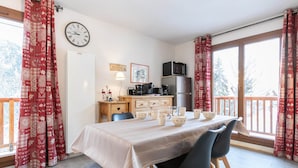 Un comedor con una mesa dispuesta para cuatro, una cocina con electrodomésticos y un balcón con cortinas rojas.