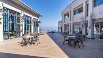 An outdoor patio area with tables and chairs, large windows, and a view of the sea.