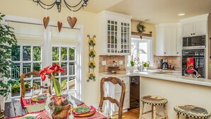 A well-lit kitchen with white cabinets, a dining table set with red and green tableware, and a view of the outdoors through large windows.