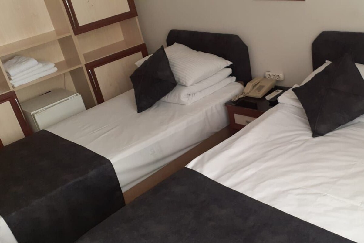 A hotel room with two beds, each with white linens and dark pillows. There is a bedside table with a phone and a lamp.