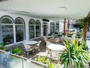 A covered outdoor seating area with multiple tables and chairs, surrounded by potted plants and large windows. - LEMAN AKÇAY OTEL (Edremit)