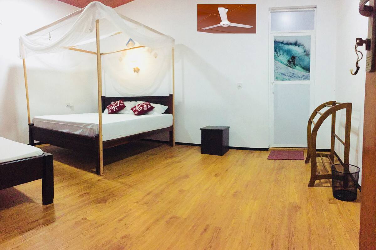 Une chambre avec un lit à baldaquin, une petite table de chevet, un plancher en bois, une porte ornée d’une affiche et un ventilateur de plafond.