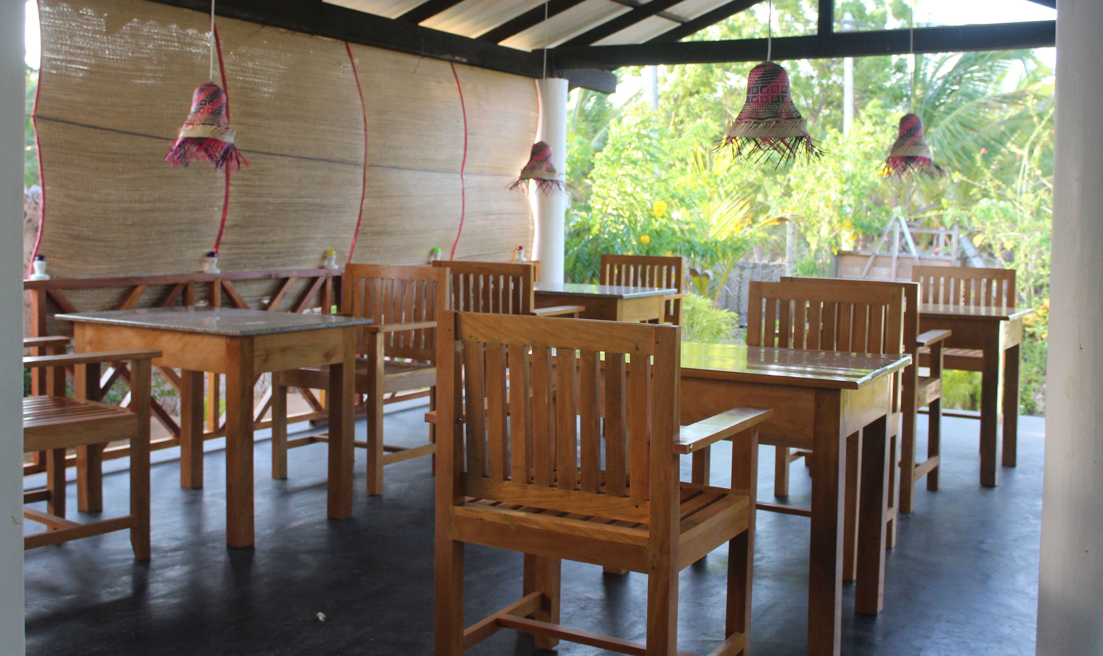 A covered outdoor seating area with wooden tables and chairs, a woven wall, and hanging decorations.