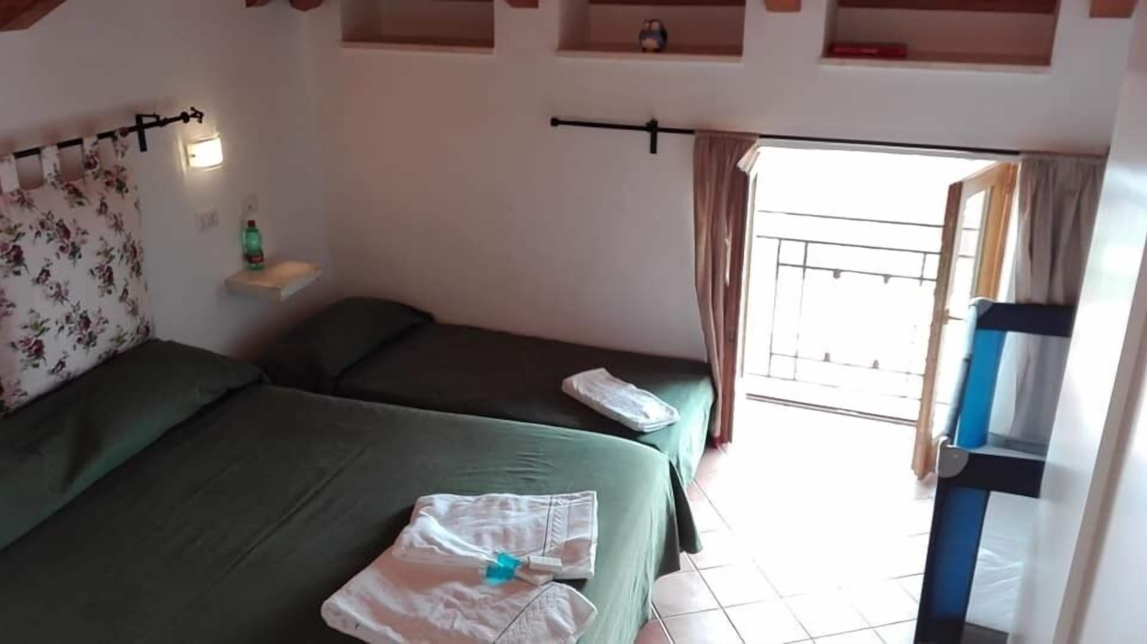 A bedroom with two beds, a small shelf with a bottle, and a window with curtains.