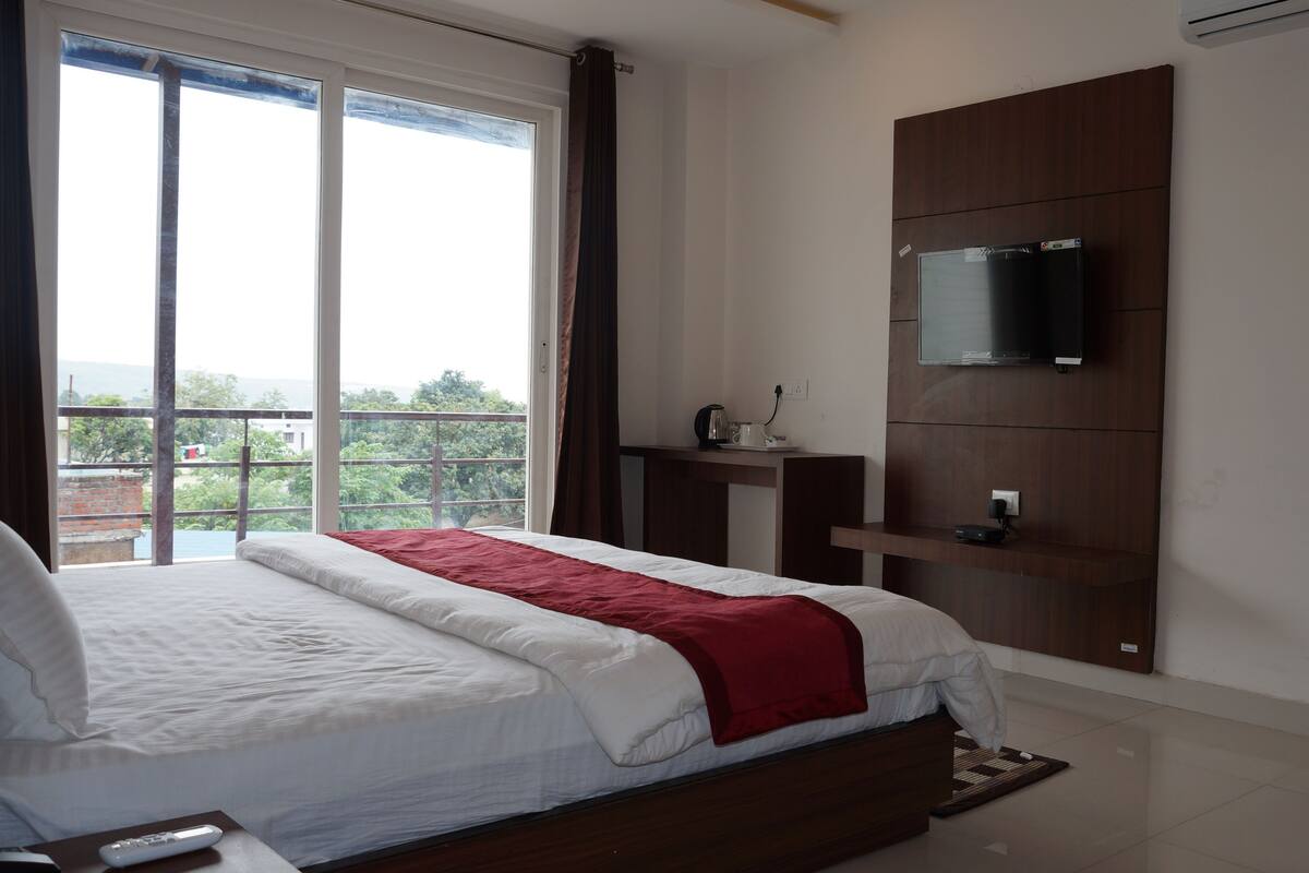 A bedroom with a large bed, a TV mounted on a wooden panel, a small table with a kettle, and a view of greenery through the window.
