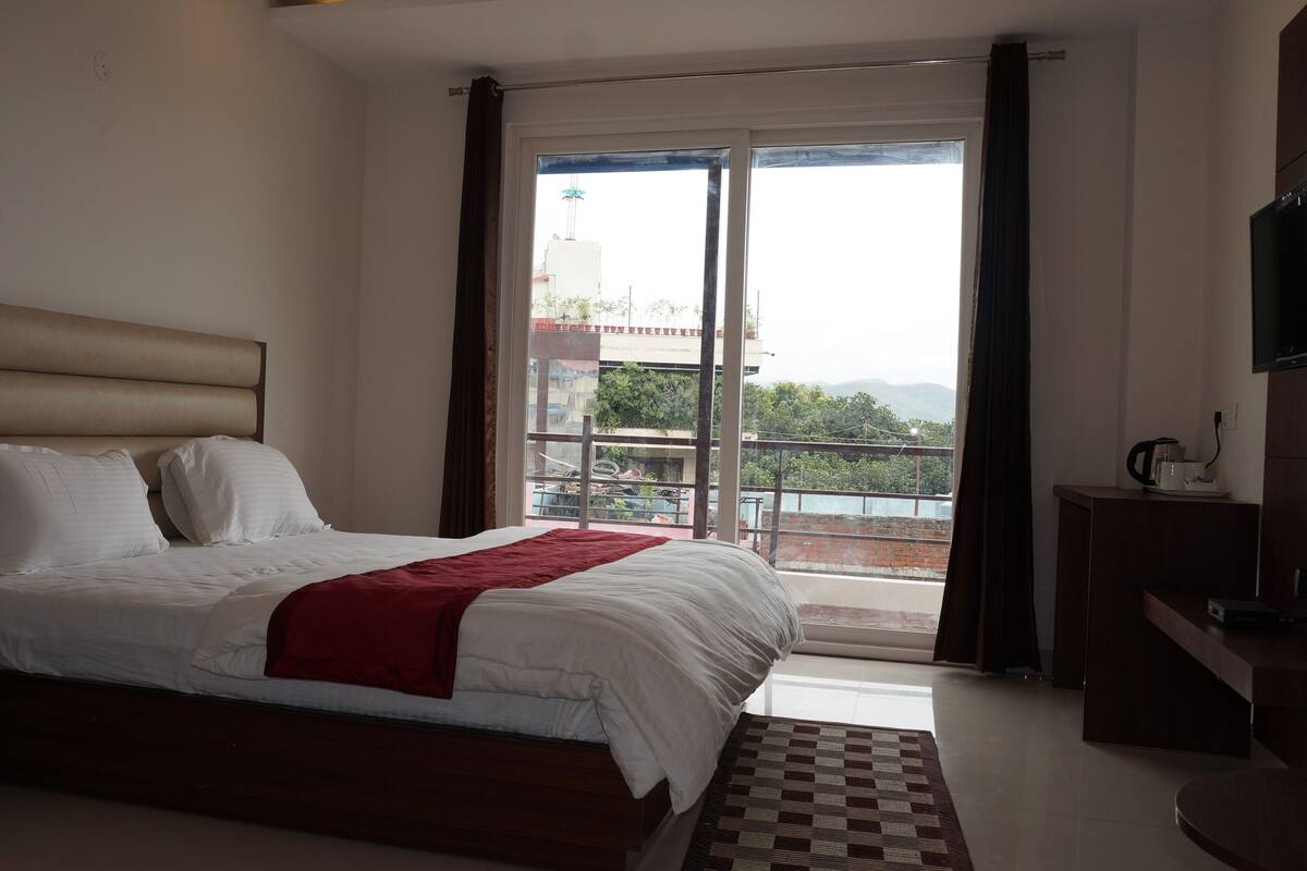 A hotel room with a bed, a bedside table, a TV, and a balcony with a view.