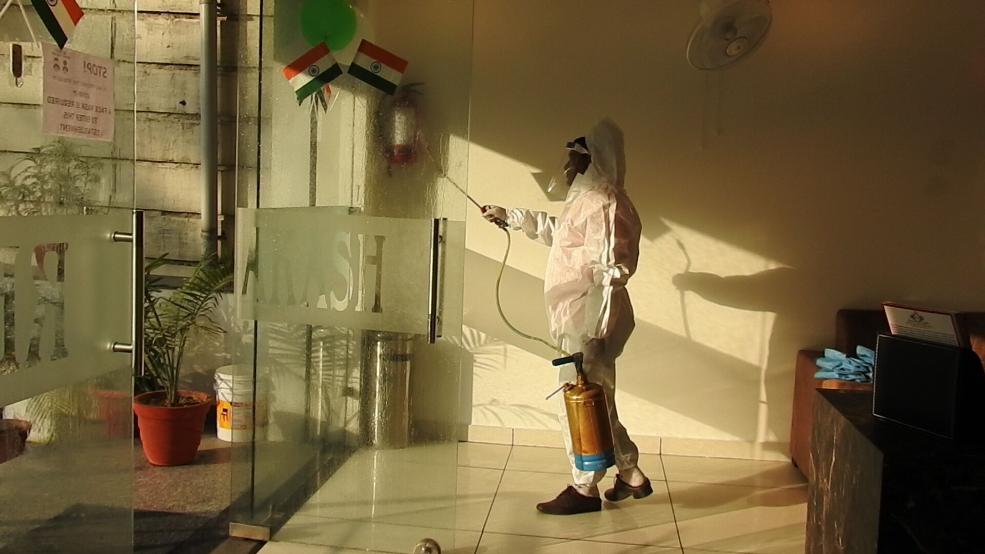 Una persona con traje de protección rociando una superficie en una habitación con puerta de cristal, una planta en maceta y un cartel en la pared.