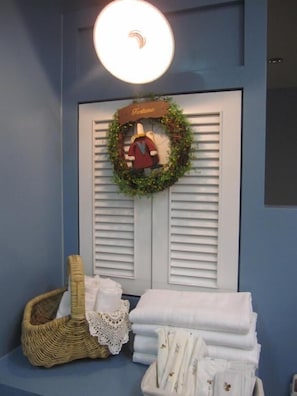 A room with a blue wall, a white door with shutters, a lit ceiling light, a wreath with a figure, a wicker basket, and folded towels. - Sweet Home 101 (Guangfu)