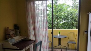 Una habitación con cama, escritorio, silla, una mesita con planta, y un balcón con sillas y mesa.