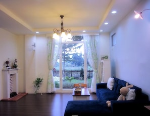 A living room with a dark blue sofa, a chandelier, a fireplace, and a view of a garden through large windows. - Sweet Home 101 (Guangfu)