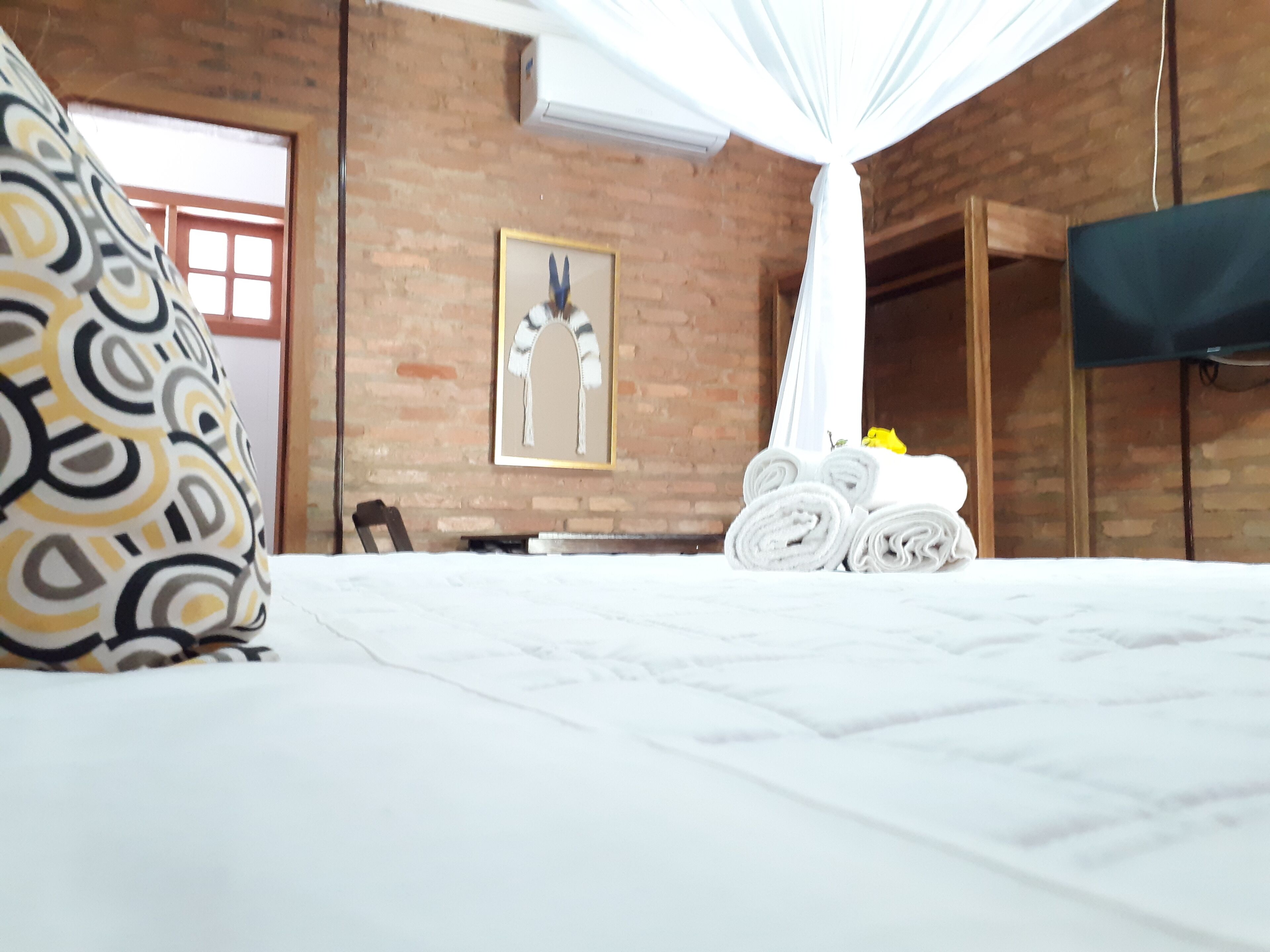 Una habitación de hotel con una cama, dosel blanco, pared de ladrillo, un cuadro enmarcado y un televisor montado en la pared.