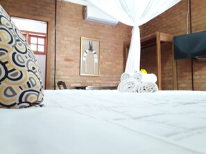 Una habitación de hotel con una cama, dosel blanco, pared de ladrillo, un cuadro enmarcado y un televisor montado en la pared.