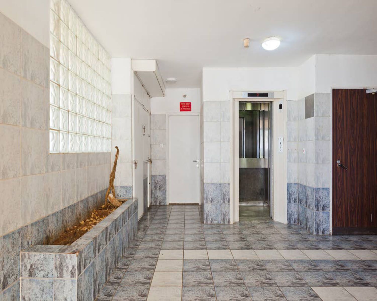 A hallway with a tiled floor, a wooden door, a glass partition, and an elevator.