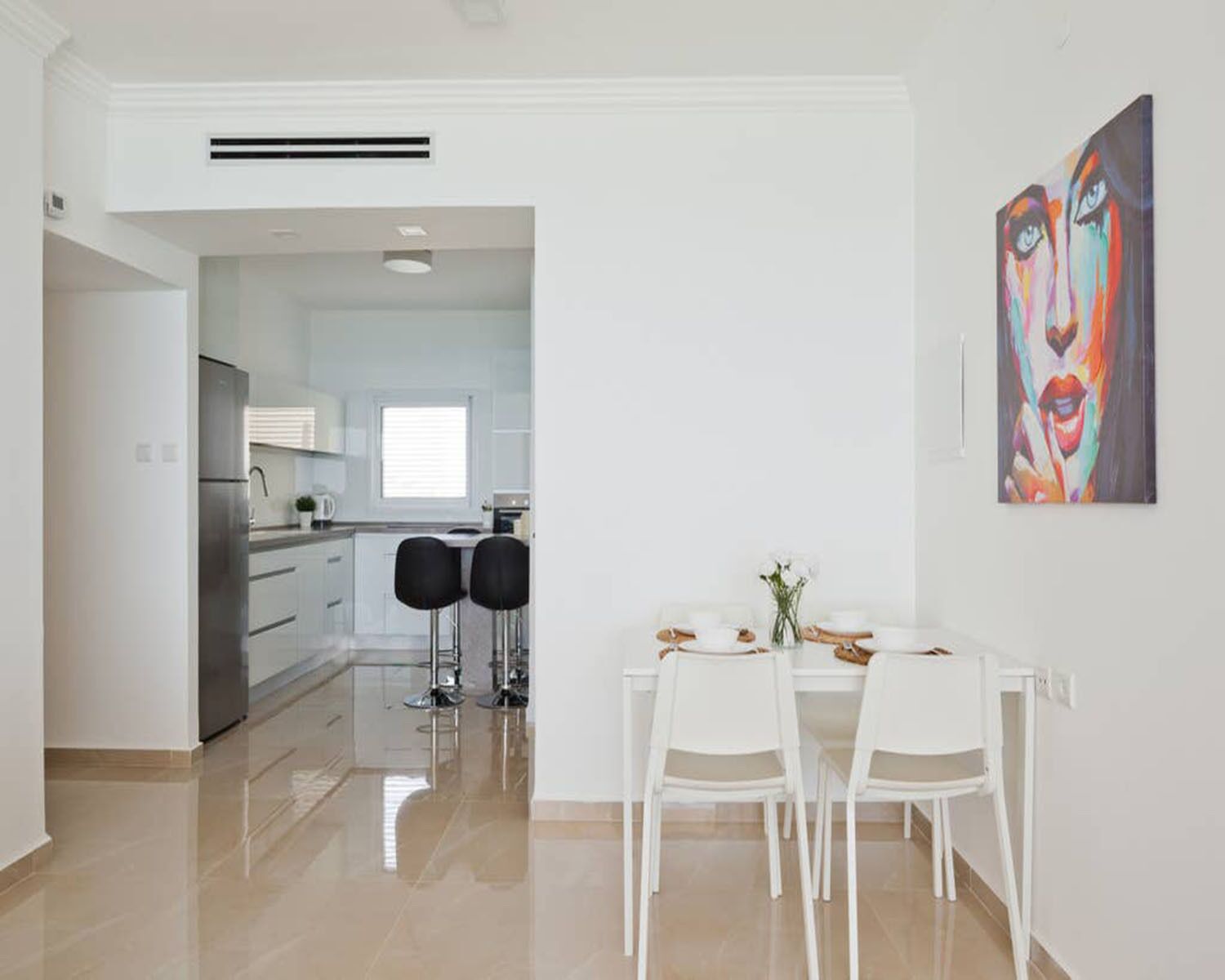 A modern kitchen with white cabinets and stainless steel appliances, a dining area with white chairs and a table, and a painting on the wall.