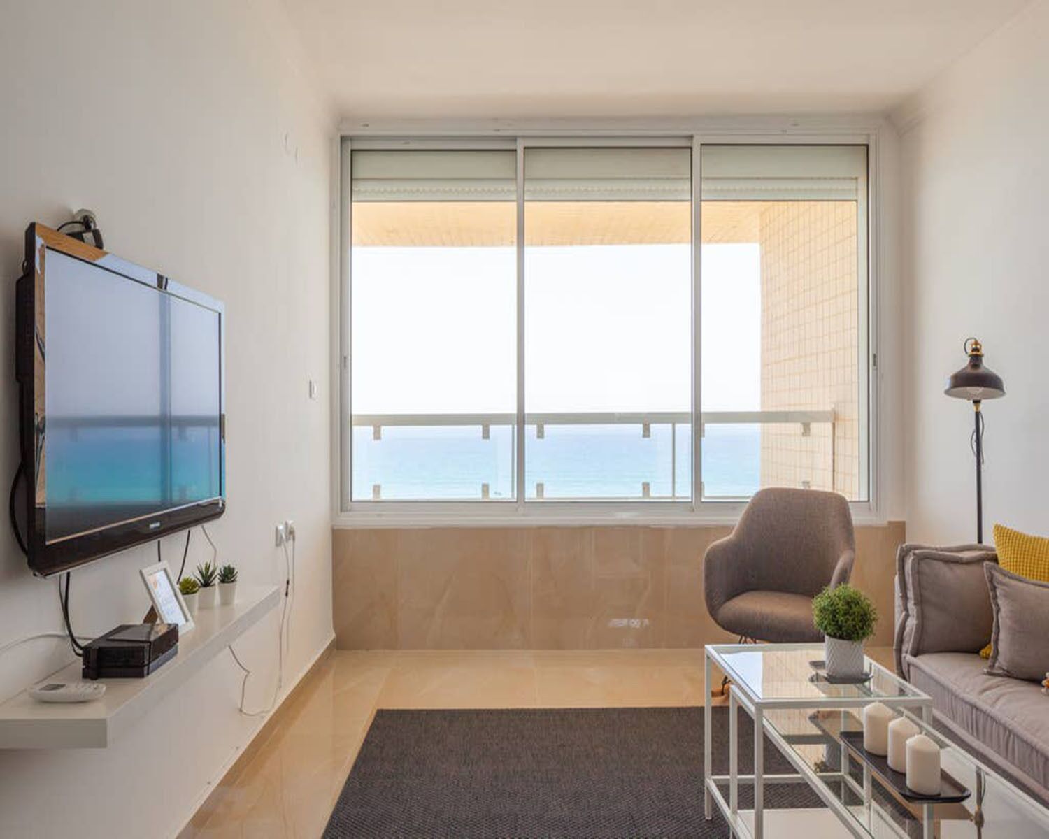 A modern living room with a large flat-screen TV, a comfortable sofa, a glass coffee table, and a view of the sea through a large window.