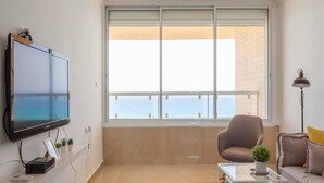 A modern living room with a large flat-screen TV, a comfortable sofa, a glass coffee table, and a view of the sea through a large window. - 3BR Apt Beach View Best Loc Bat-Yam (Bat Yam)
