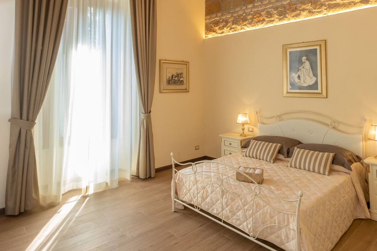 Une chambre à coucher avec un grand lit, un lustre, un mur en briques et une fenêtre avec des rideaux.