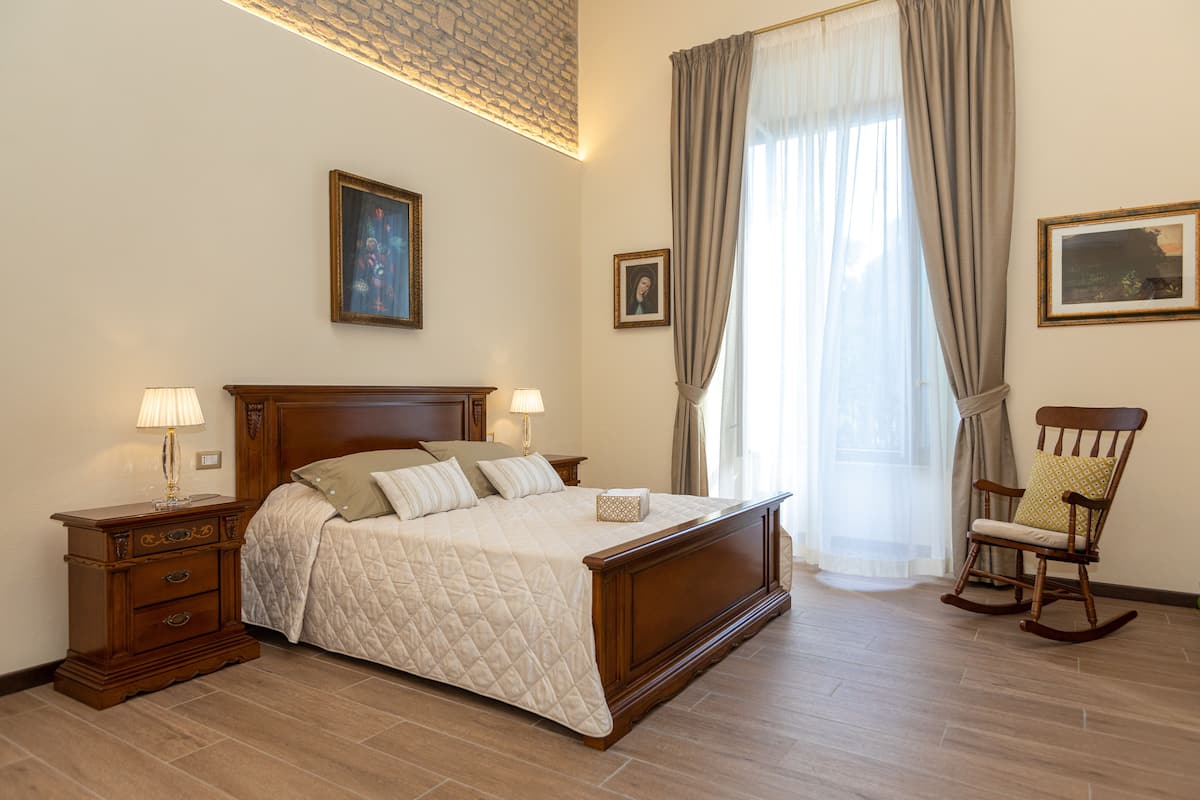 Une chambre à coucher avec un grand lit, deux tables de chevet avec des lampes, un fauteuil à bascule et une fenêtre avec des rideaux.