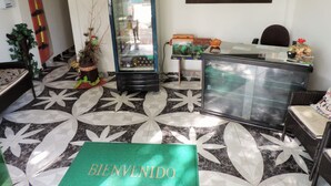 A reception area with a 'BIENVENIDO' welcome mat, a glass display case with decorative items, a wall-mounted abstract painting, and a patterned floor.