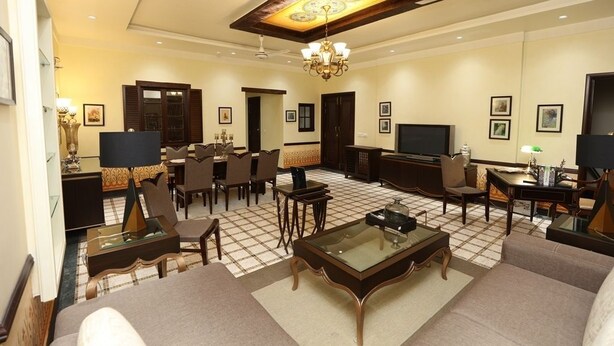 A spacious living room with a large sofa, a coffee table, and a TV. There are multiple chairs and a dining table with chairs. The room has a high ceiling with a chandelier and recessed lighting.
