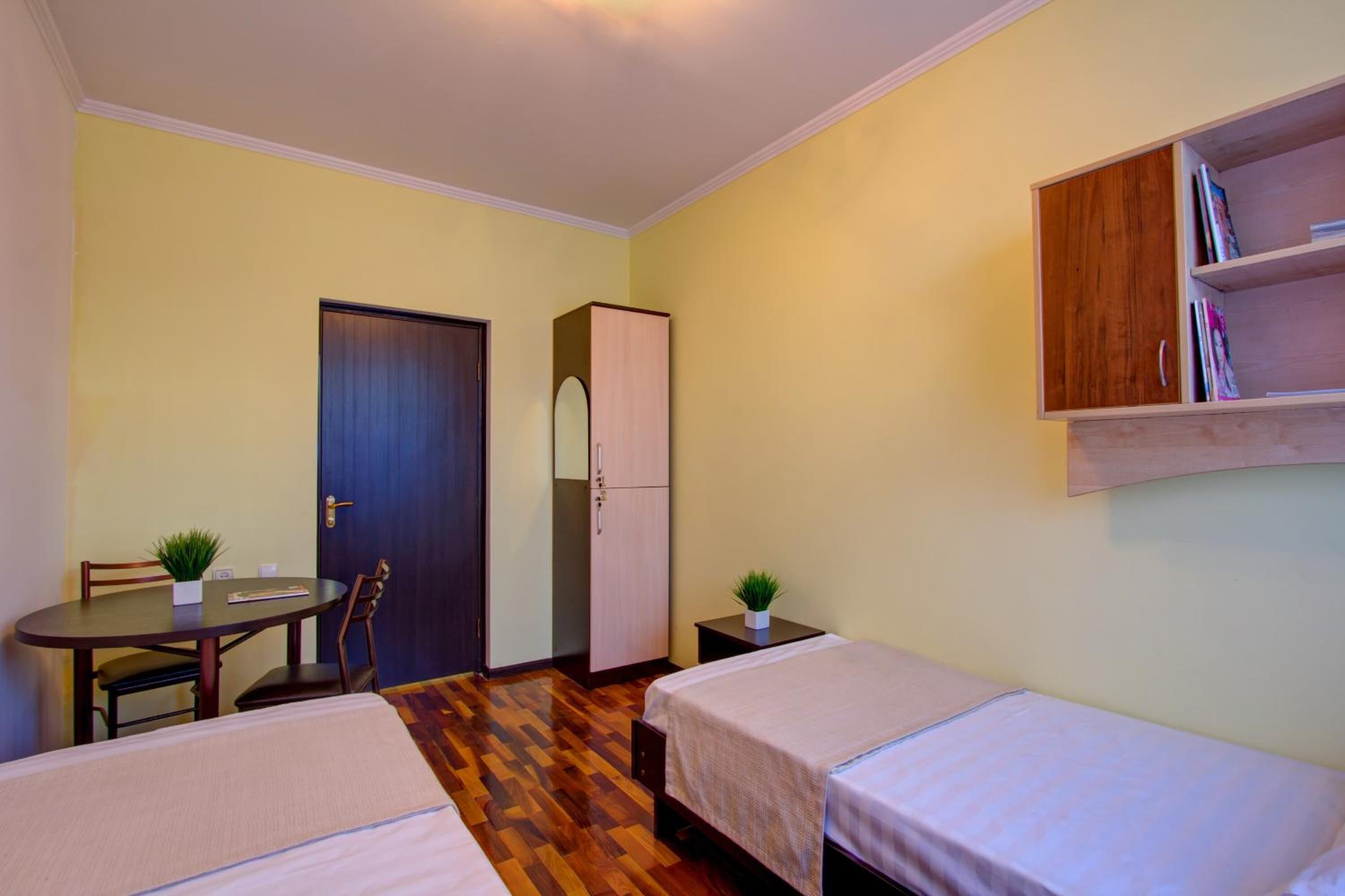 Una habitación pequeña y luminosa con dos camas individuales, un armario de madera, una mesita con sillas y una estantería.