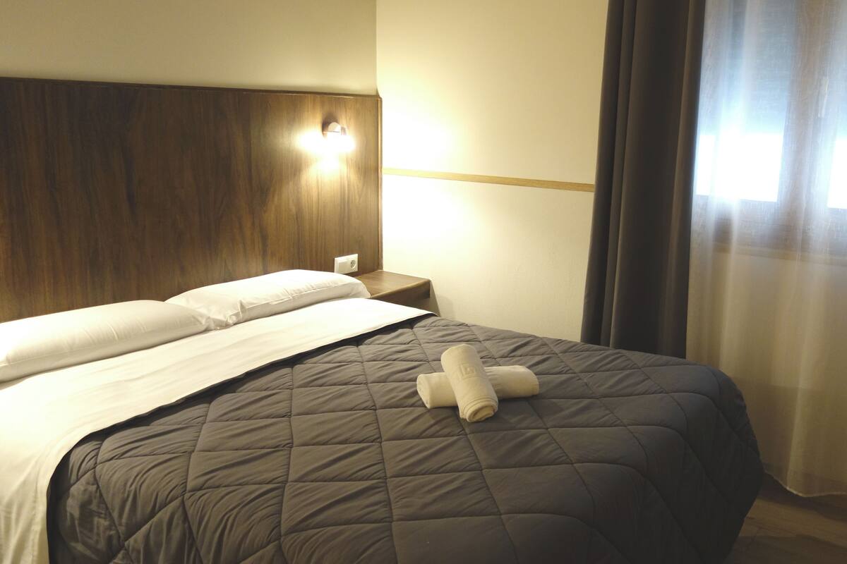 Habitación de hotel con una cama grande, cabecera de madera, mesita de noche y ventana con cortinas.
