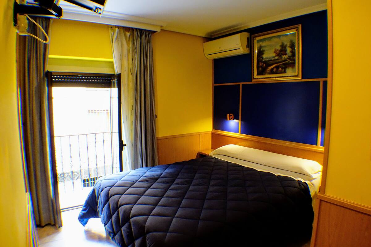 Habitación de hotel con cama, ventana con cortinas, unidad de aire acondicionado y un cuadro en la pared.