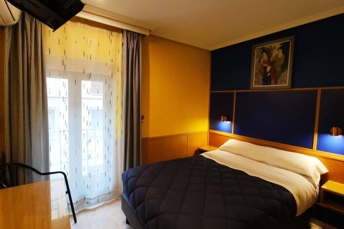Habitación de hotel con cama, escritorio y silla. Se ve una ventana con cortinas y un cuadro en la pared.