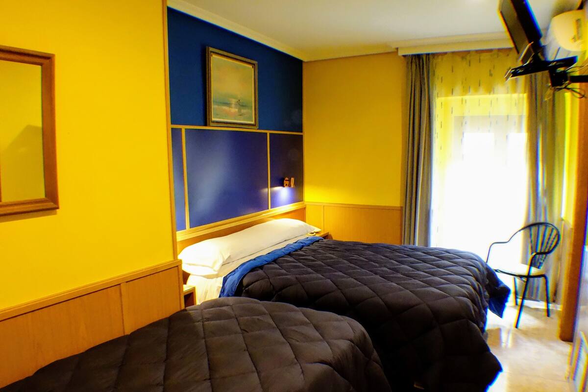 Habitación de hotel con dos camas, una pared azul decorativa, un cuadro y una silla junto a la ventana.