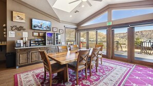 A spacious room with a wooden dining table, chairs, a ceiling fan, a TV, and a large window offering a view of the desert. - Zion Cliff Lodge (Hildale)