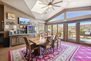 A spacious room with a large dining table, chairs, a ceiling fan, a kitchen area with a coffee station, and large windows offering a view of the desert landscape. - Zion Cliff Lodge (Hildale)
