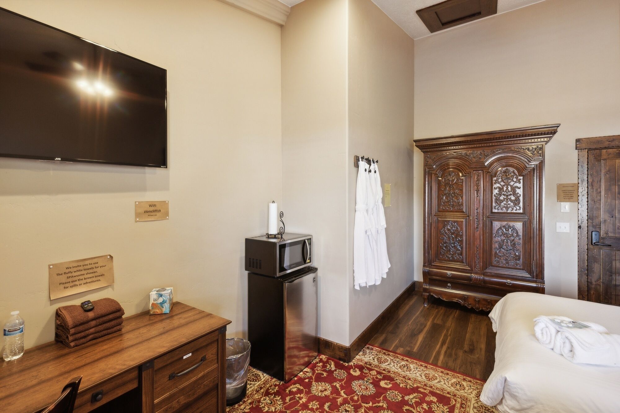 A hotel room with a flat-screen TV, a wooden dresser, a microwave, a red patterned rug, a bed with white linens, and an ornate wooden wardrobe.