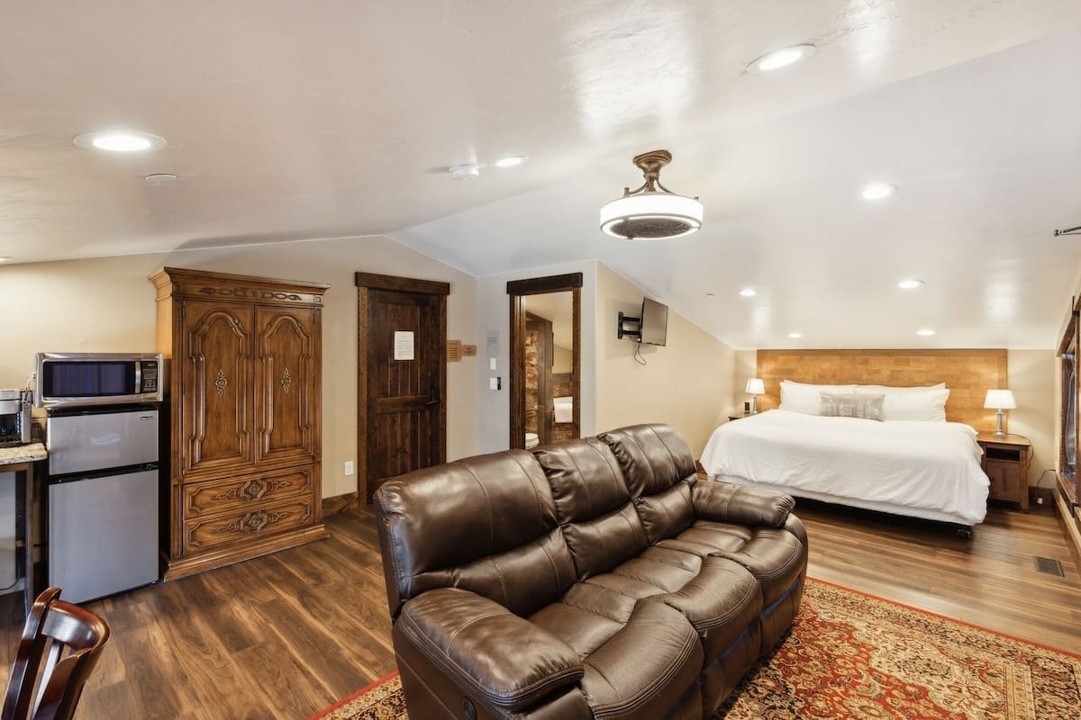 A hotel room with a bed, a leather sofa, a wooden wardrobe, a kitchenette, and a ceiling fan.