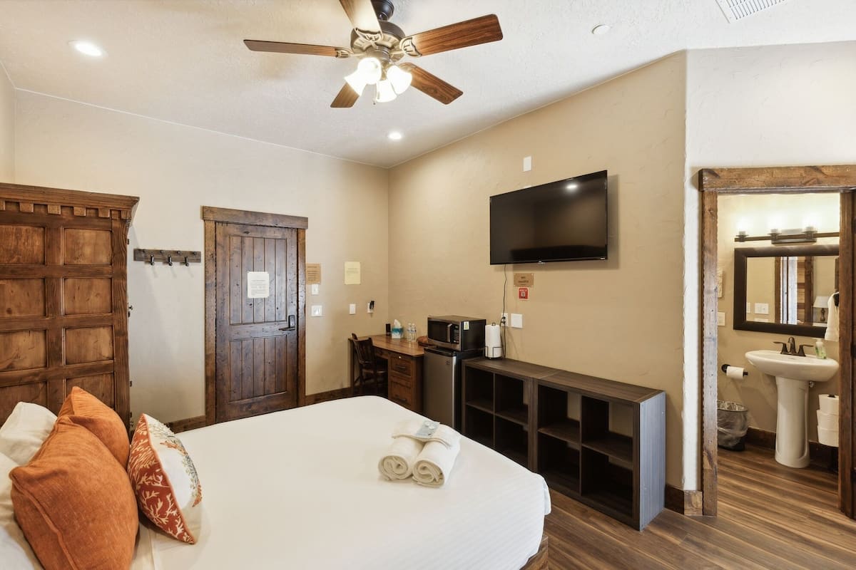 A hotel room with a large bed, a ceiling fan, a flat-screen TV, a wooden door, a small kitchenette, and a bathroom with a sink and mirror.