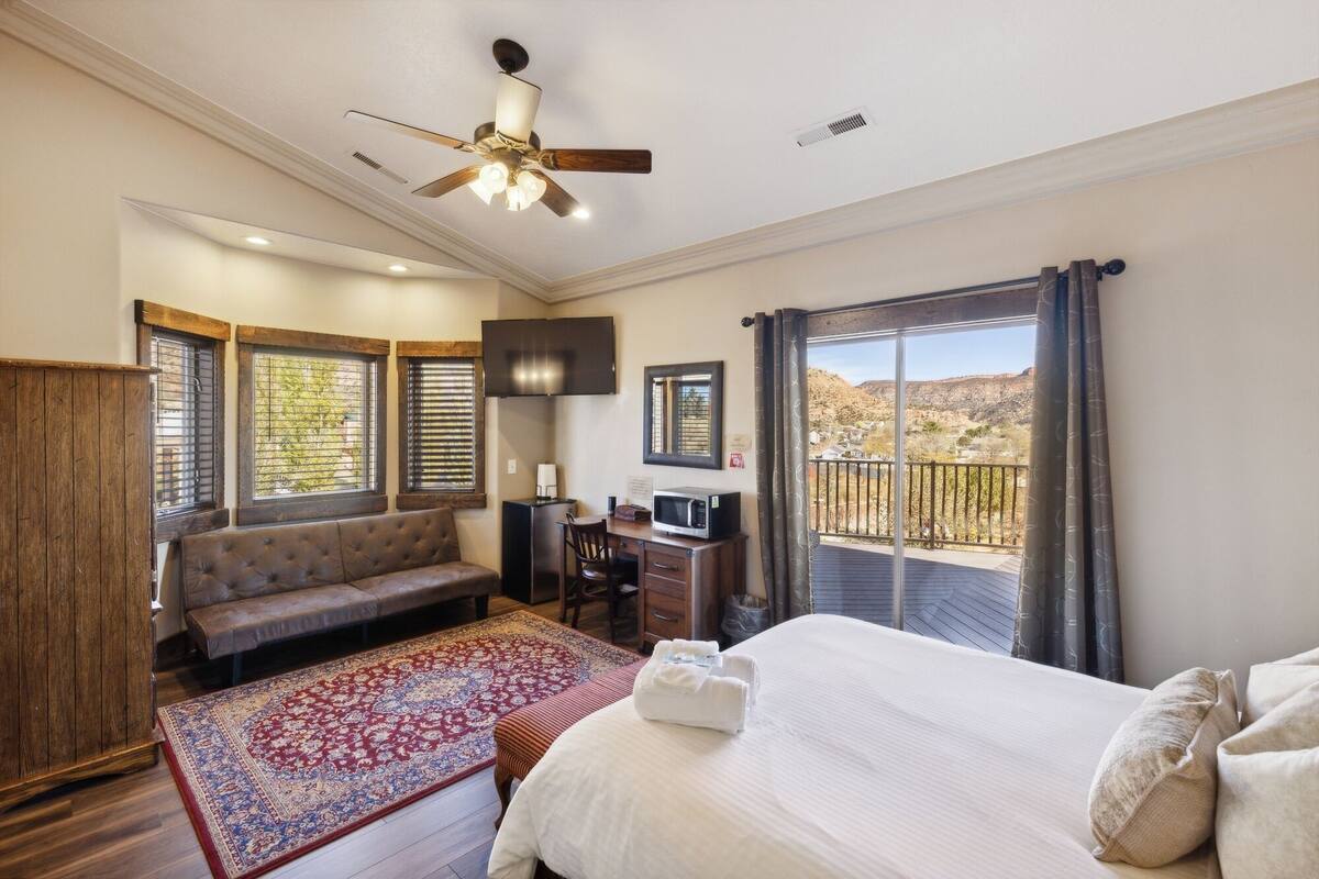 A bedroom with a bed, a sofa, a desk, a ceiling fan, and a view of the outdoors.