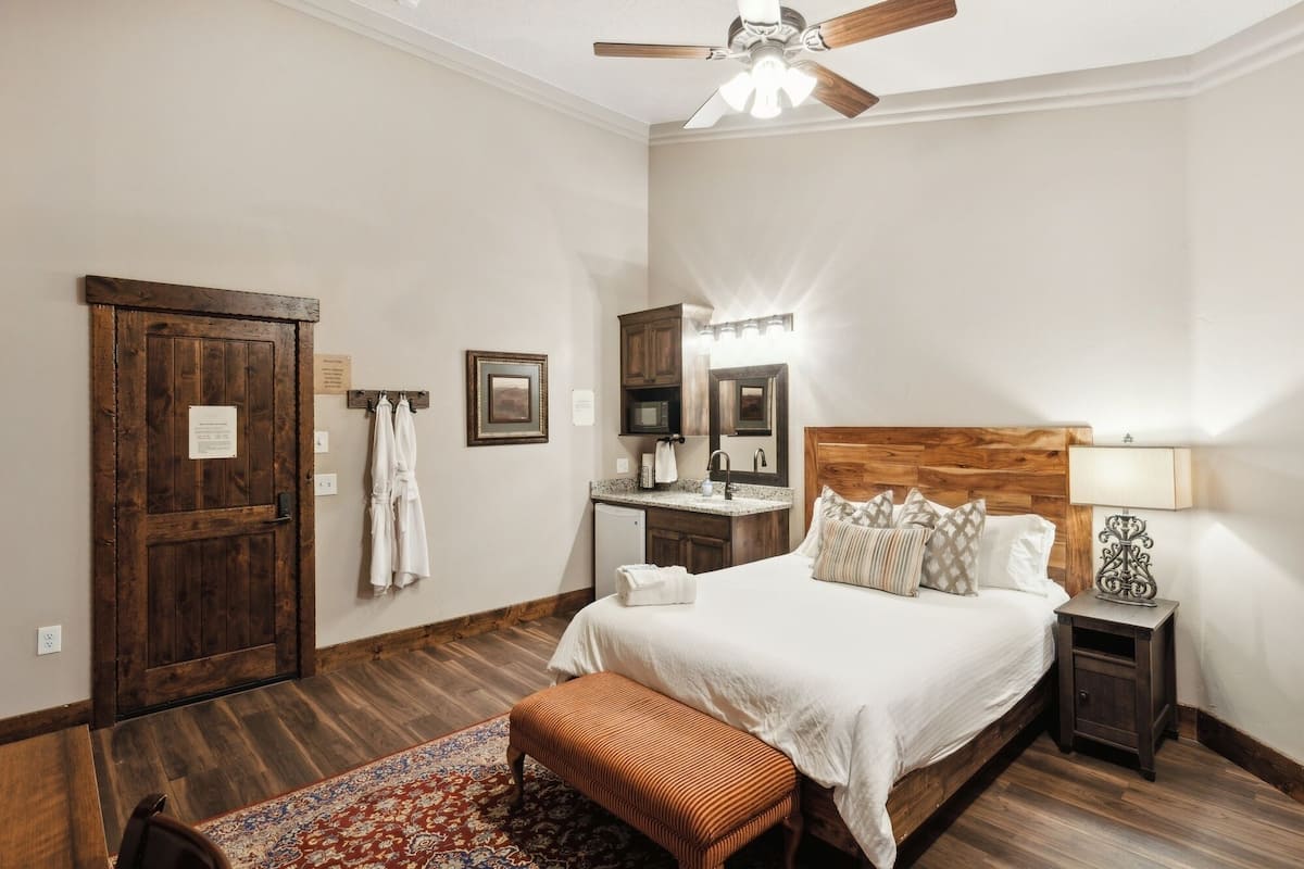 A bedroom with a wooden headboard, a bed with white linens, a nightstand with a lamp, a wooden floor, a ceiling fan, and a bathroom with a wooden door and towel rack.
