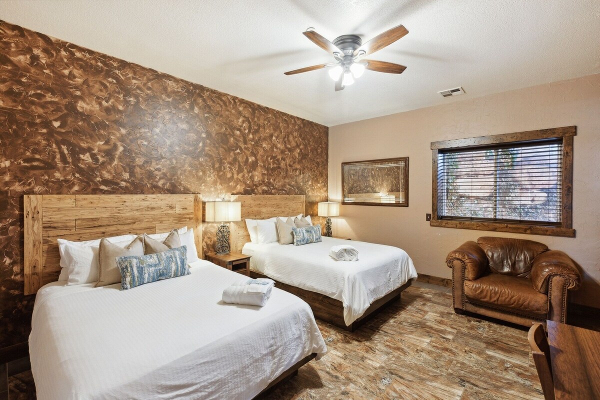 A hotel room with two beds, a ceiling fan, a window with blinds, and a wooden floor.