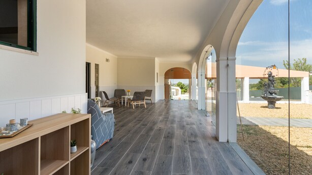 Amplio espacio interior con piso de madera, zona de estar con sillas y una mesa, y vista a un jardín exterior a través de un ventanal.