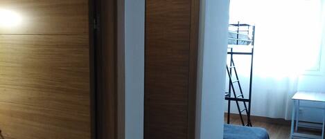 A room with a wooden wardrobe, a bed with a white mattress, a small table, and a window with curtains.